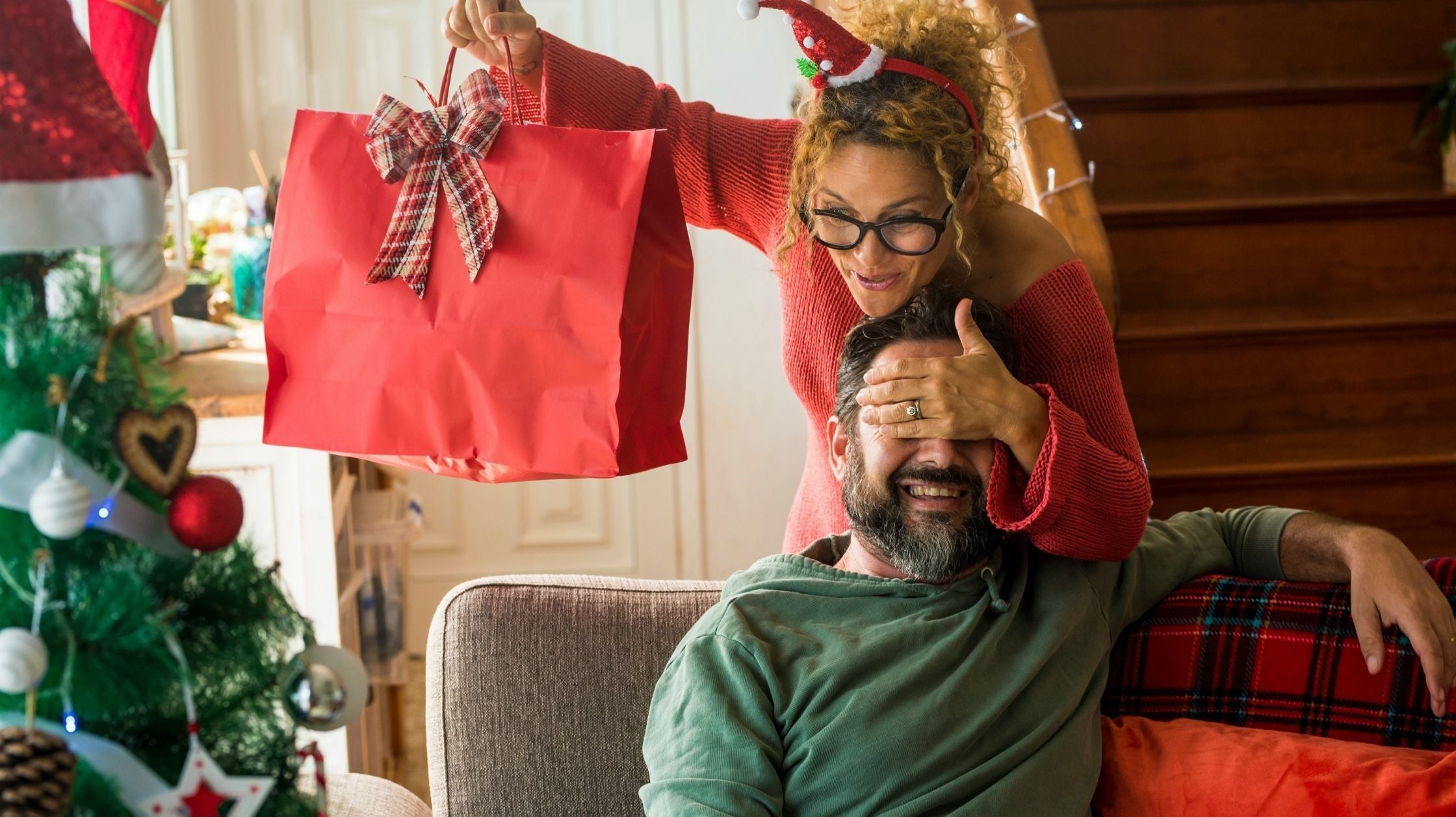 Frau überrascht lachenden Mann im weihnachtlich geschmückten Wohnzimmer mit roter Geschenktüte – Trends und Ideen zu Weihnachten 2025