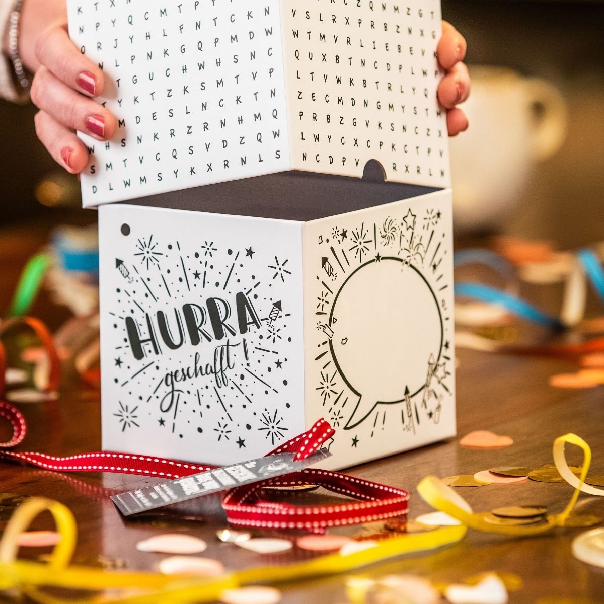 Geöffnete Rätsel-Geschenkbox zeigt Aufdruck ‚Hurra geschafft‘ – kreative Gratulationsidee